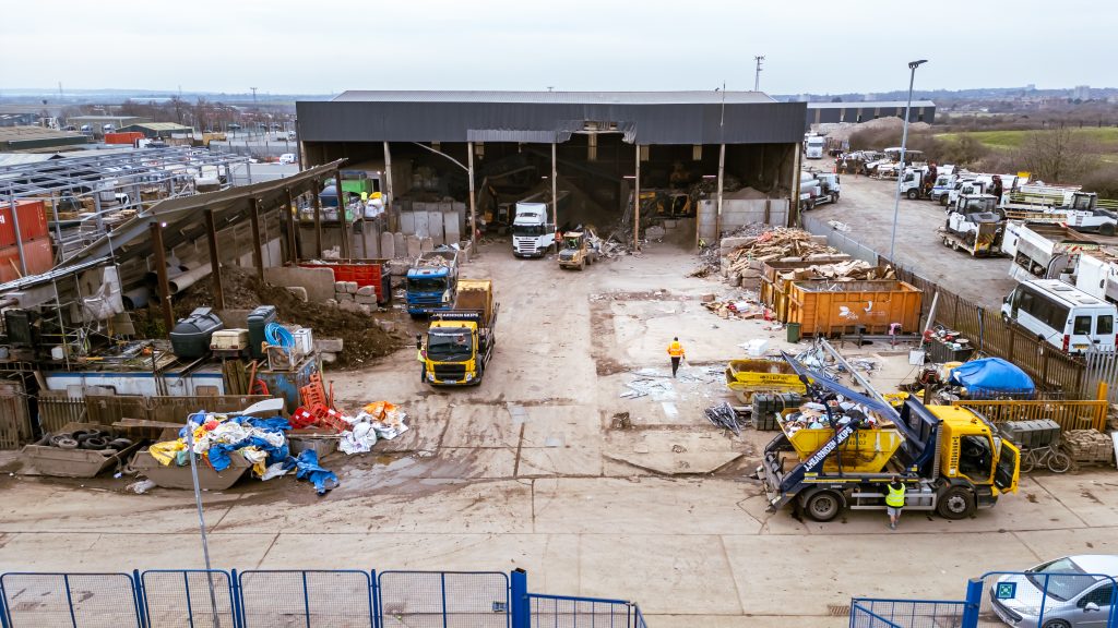 Aerial view of an industrial waste management facility with lorries, construction vehicles, waste heaps, and workers in high-visibility clothing on a concrete yard. A large shed stands in the centre.