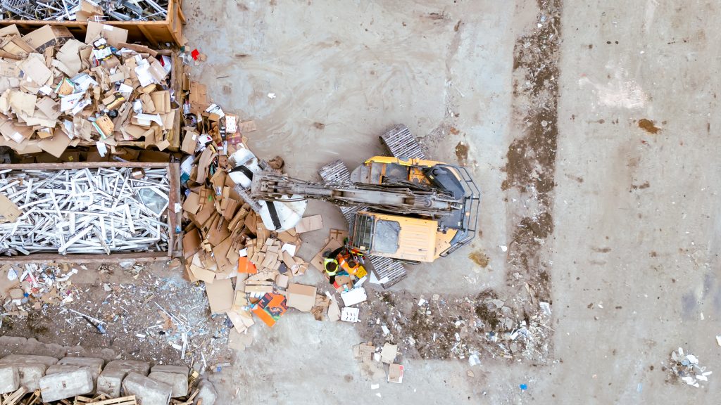Aerial view of a yellow excavator moving cardboard and metal debris at a recycling or waste facility, with scattered materials and two workers visible near the machine.
