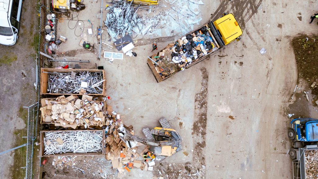 Aerial view of a recycling or waste processing facility with piles of scrap metal, cardboard, and rubbish in containers, a yellow lorry, a forklift, and scattered debris on the ground.