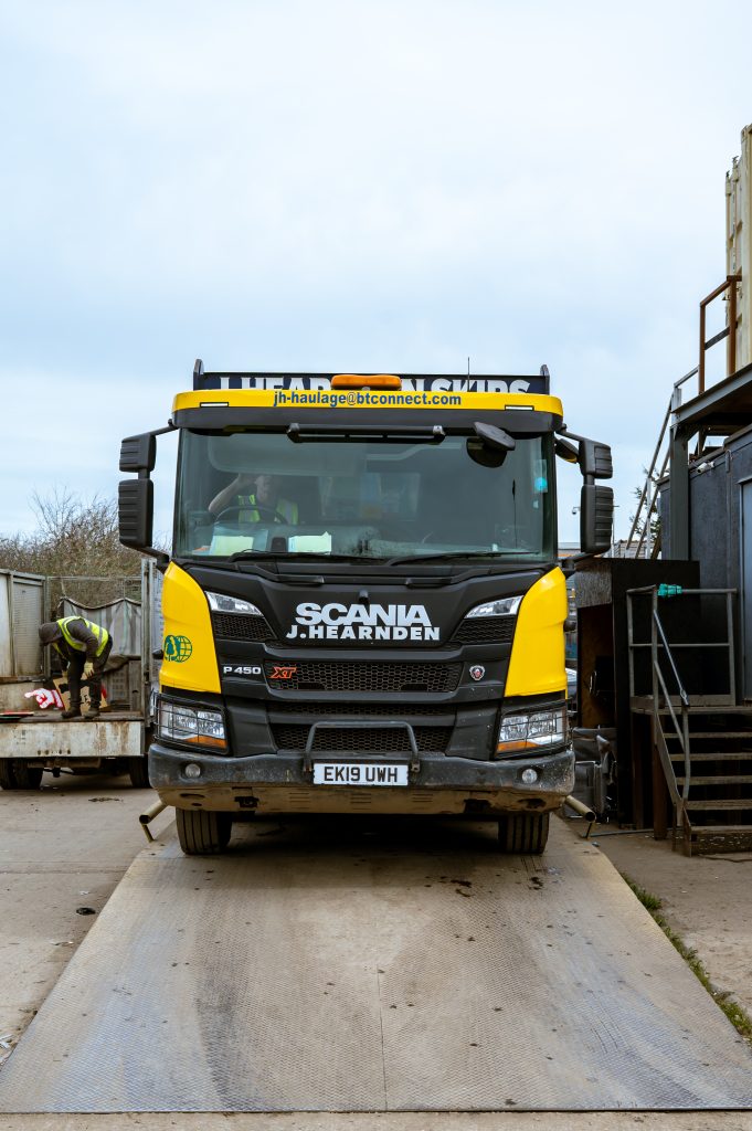 A yellow and black Scania lorry with "J. Hearnden" branding is parked on a concrete ramp at an industrial site. A worker wearing a safety vest is seen in the background near a metal container.