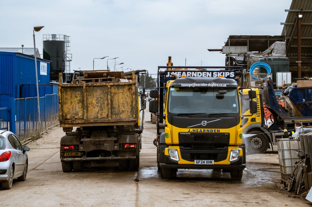 Two large yellow lorries from J Hearnden Skips are parked side by side in a busy, industrial yard with various equipment and buildings in the background. A silver car is parked on the left.