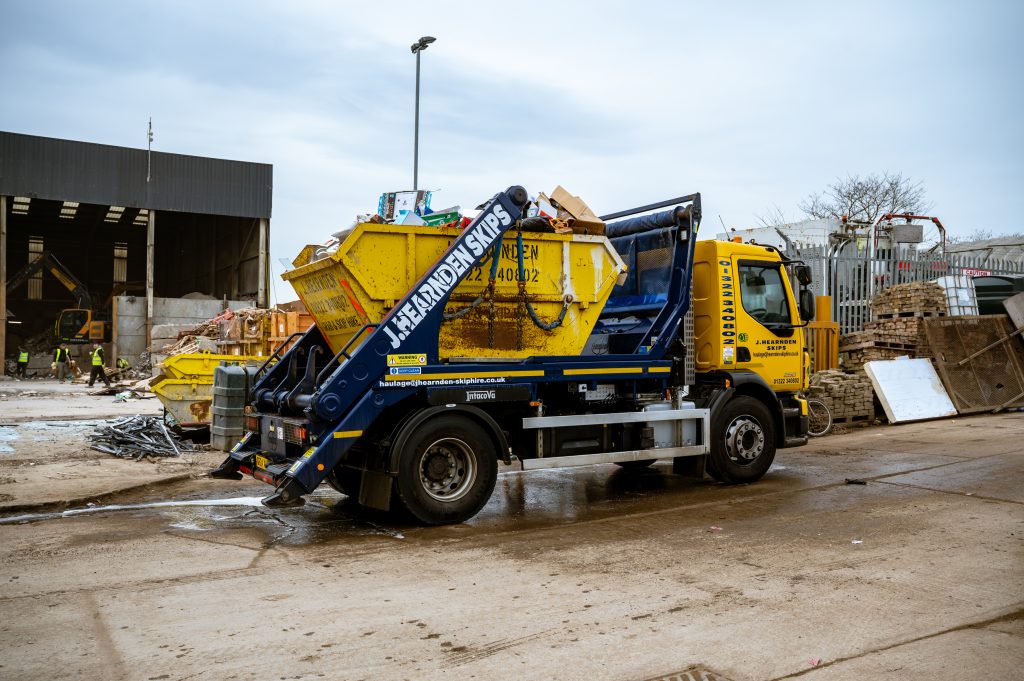 A yellow and blue skip lorry loaded with a full skip sits on a wet industrial yard, surrounded by debris, other skips, and buildings under a cloudy sky.