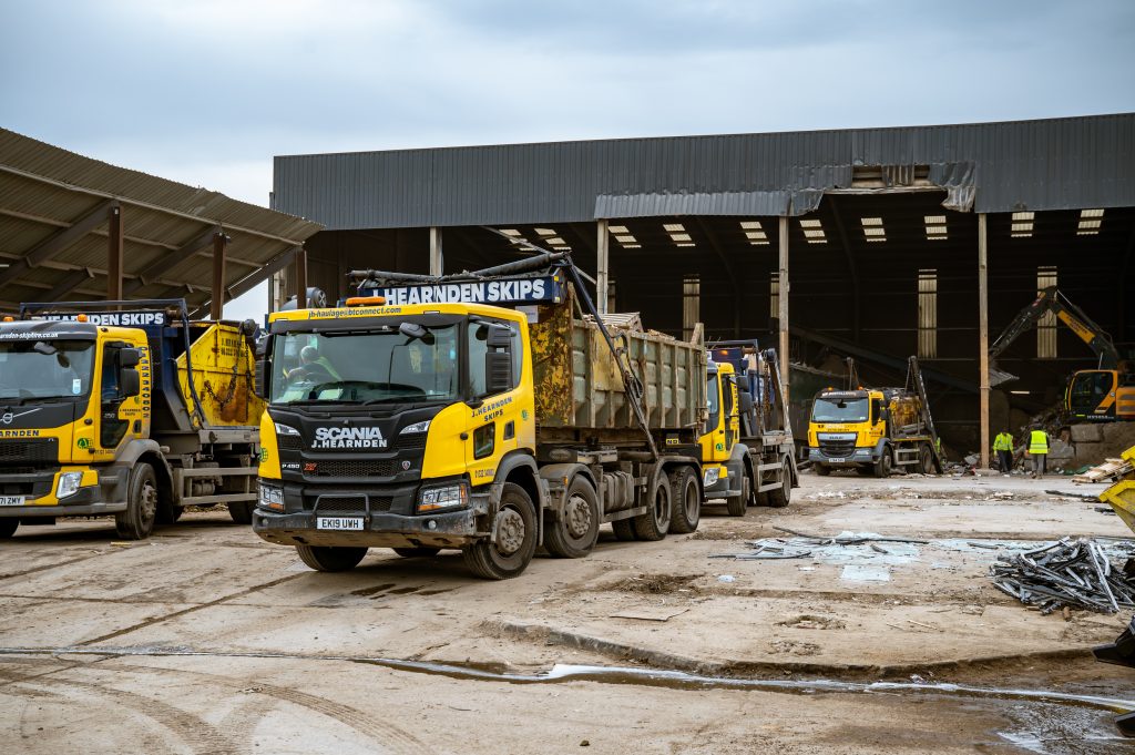 Yellow and black skip hire lorries and construction vehicles operate in a large industrial yard with a partially covered building and scattered debris under a cloudy sky.