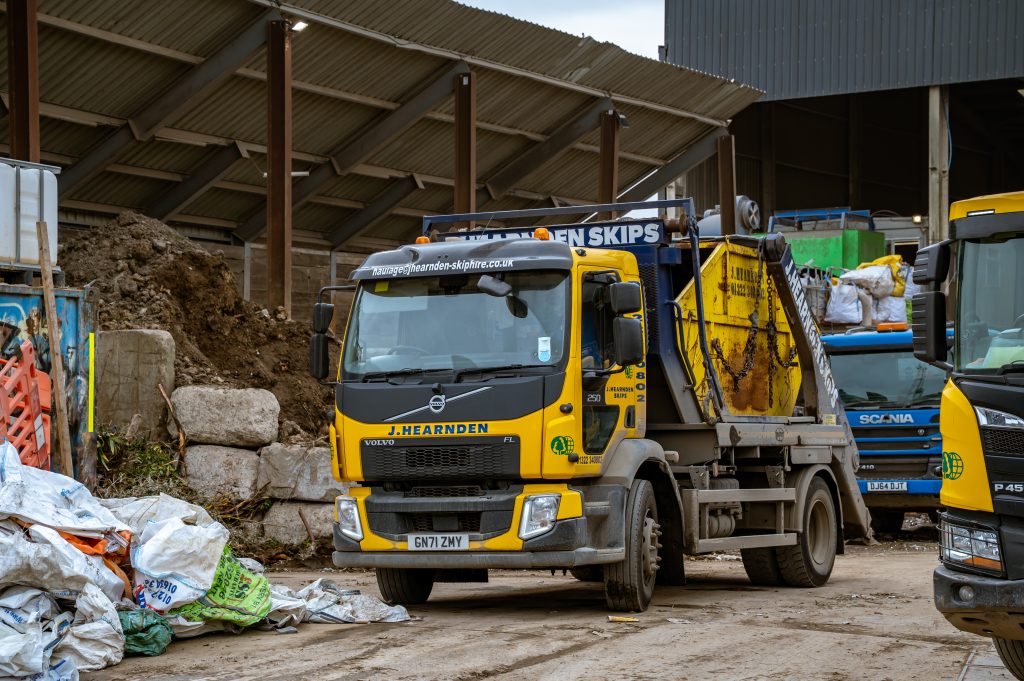 A yellow and black J. Hearnden skip lorry is parked at a waste management facility, surrounded by debris, large bags, and construction materials. Another lorry is partially visible to the right.