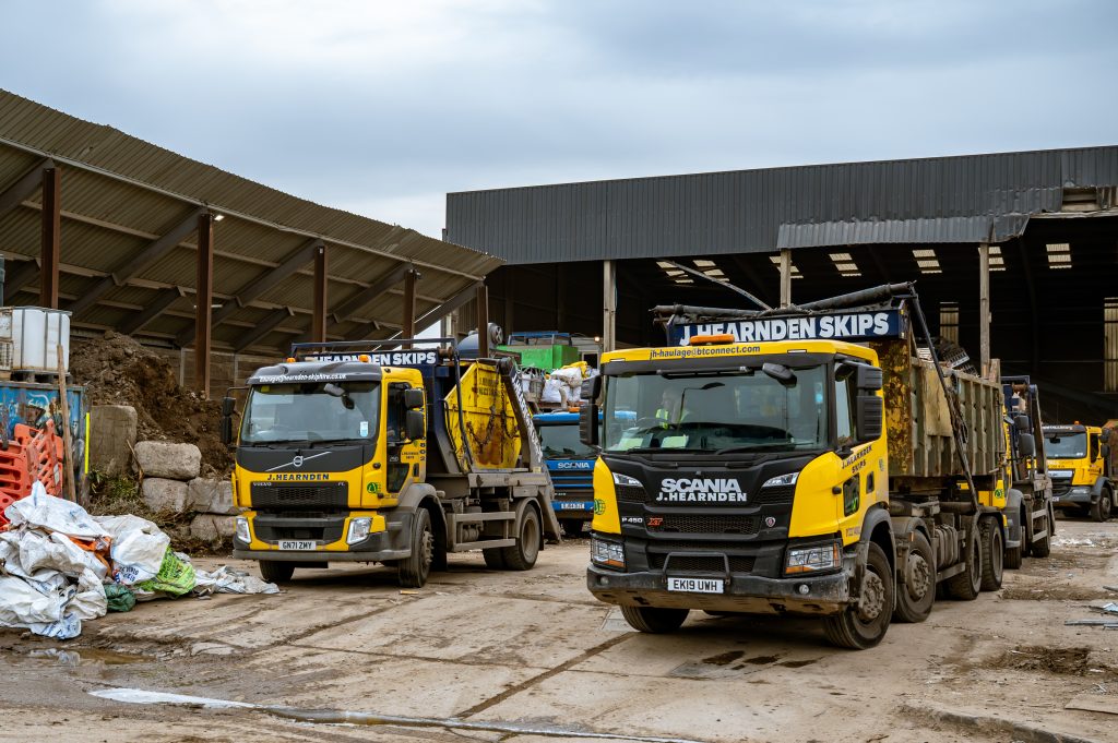 Three yellow and black skip lorries are parked in a recycling or waste management facility, with piles of materials, skips, and industrial buildings in the background. The lorries are labelled "J L FARNDEN SKIPS.