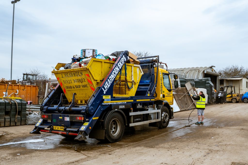 A worker in a yellow vest stands next to a blue and yellow skip lorry, which is lifting a yellow skip, in a recycling or waste management yard with various containers and buildings in the background.