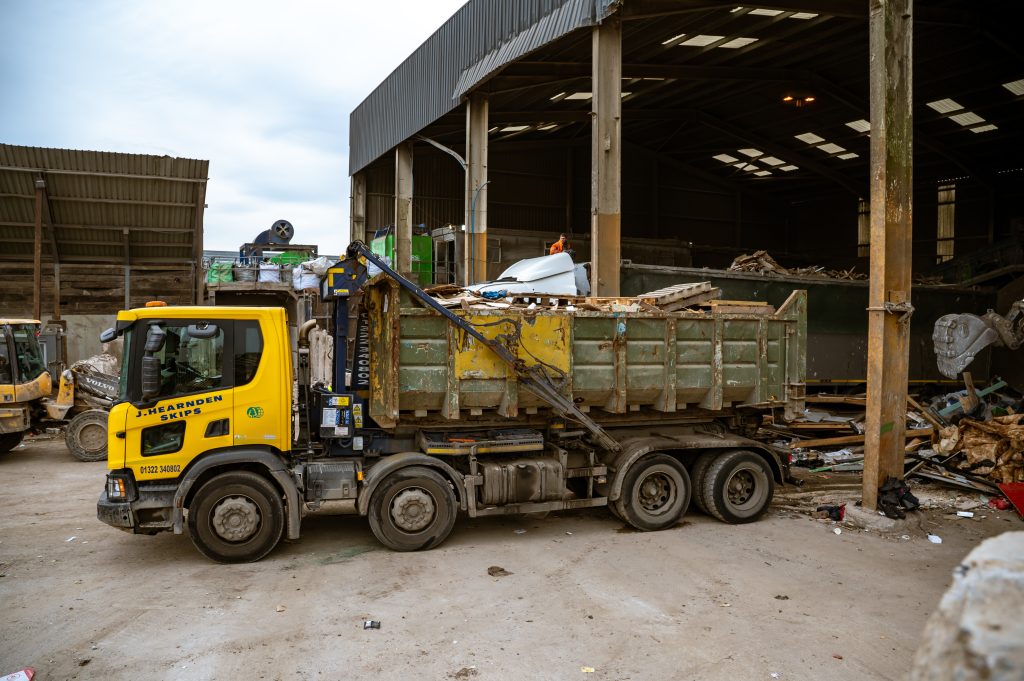 A yellow waste management lorry with a large green skip is parked outside a recycling facility, surrounded by piles of debris and construction materials.