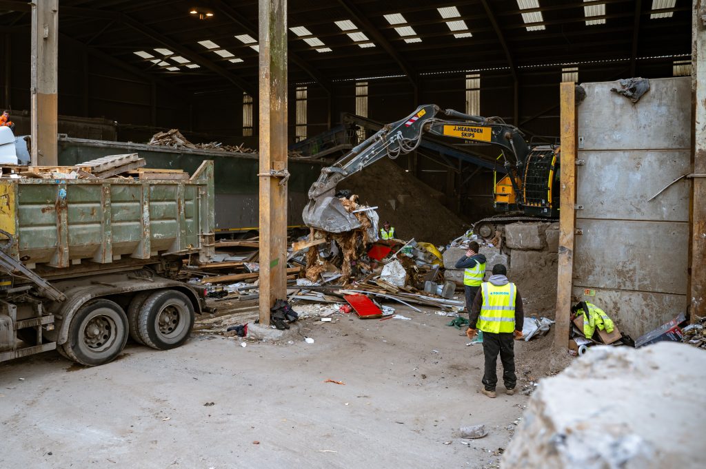 A large excavator moves debris inside a warehouse, while several workers in high-visibility jackets stand nearby. A lorry is parked on the left, and piles of mixed waste and construction materials are visible throughout the scene.