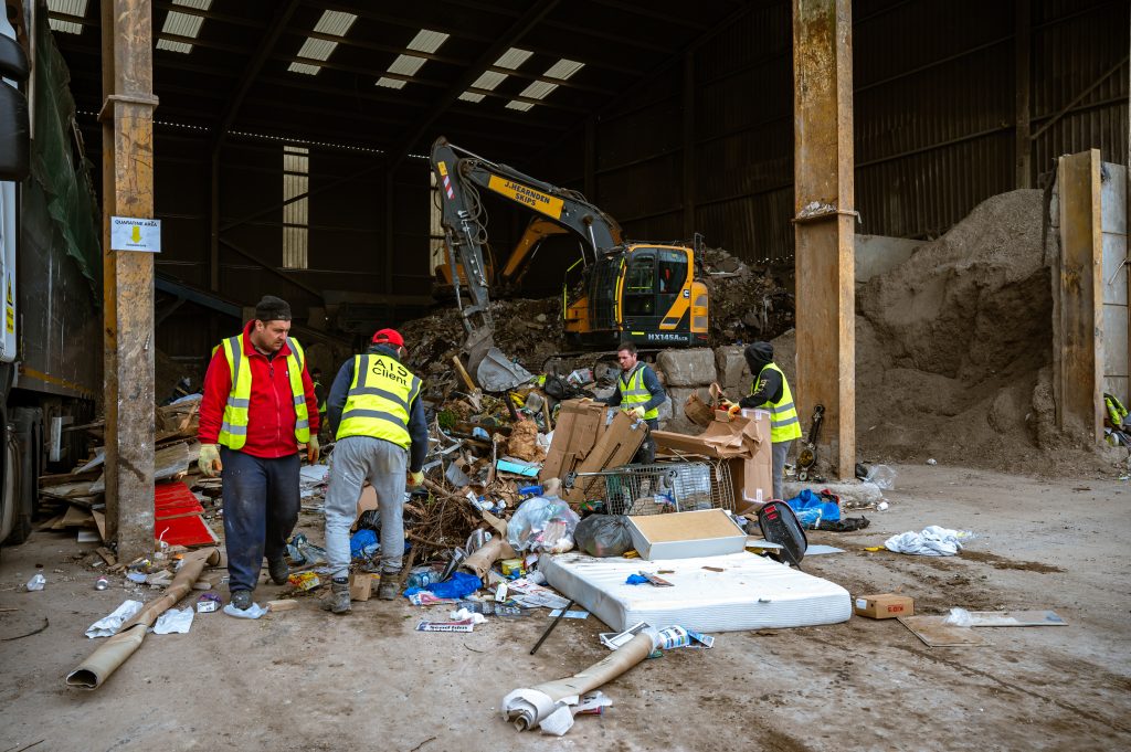 Workers in safety vests sort through piles of mixed waste, cardboard, and debris inside a large industrial warehouse, with a yellow digger and heaps of rubbish in the background.
