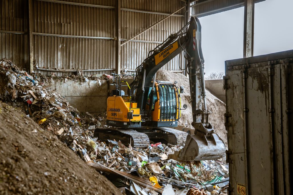 A yellow digger moves through a large indoor landfill, surrounded by piles of mixed rubbish and debris inside a warehouse-like structure.