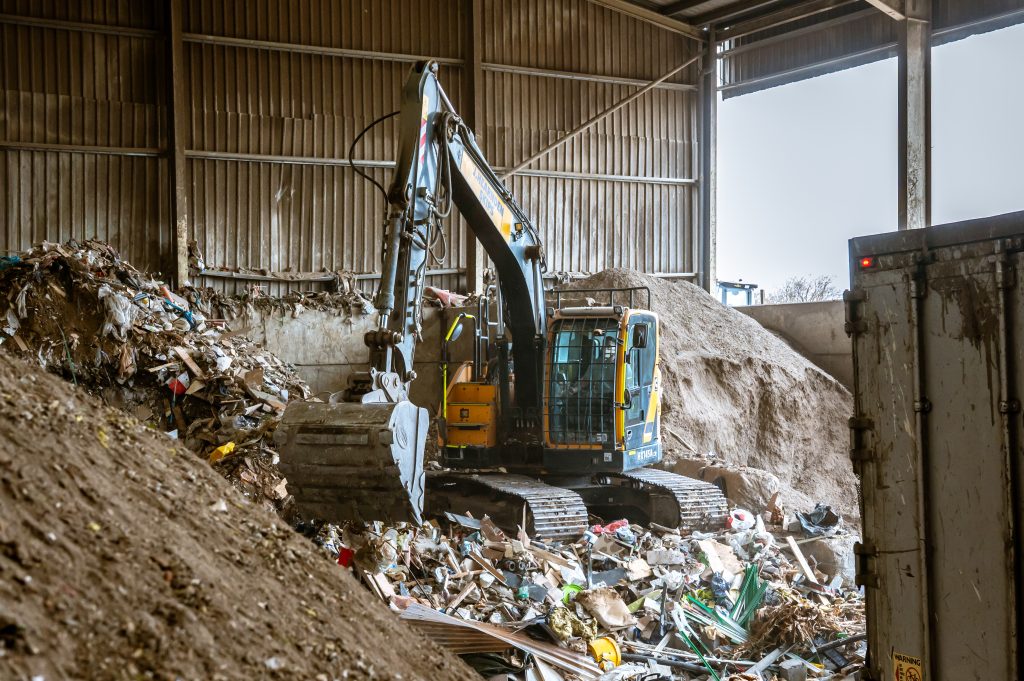 An excavator moves piles of mixed rubbish and debris inside a large industrial building, with scattered waste and a lorry visible nearby.