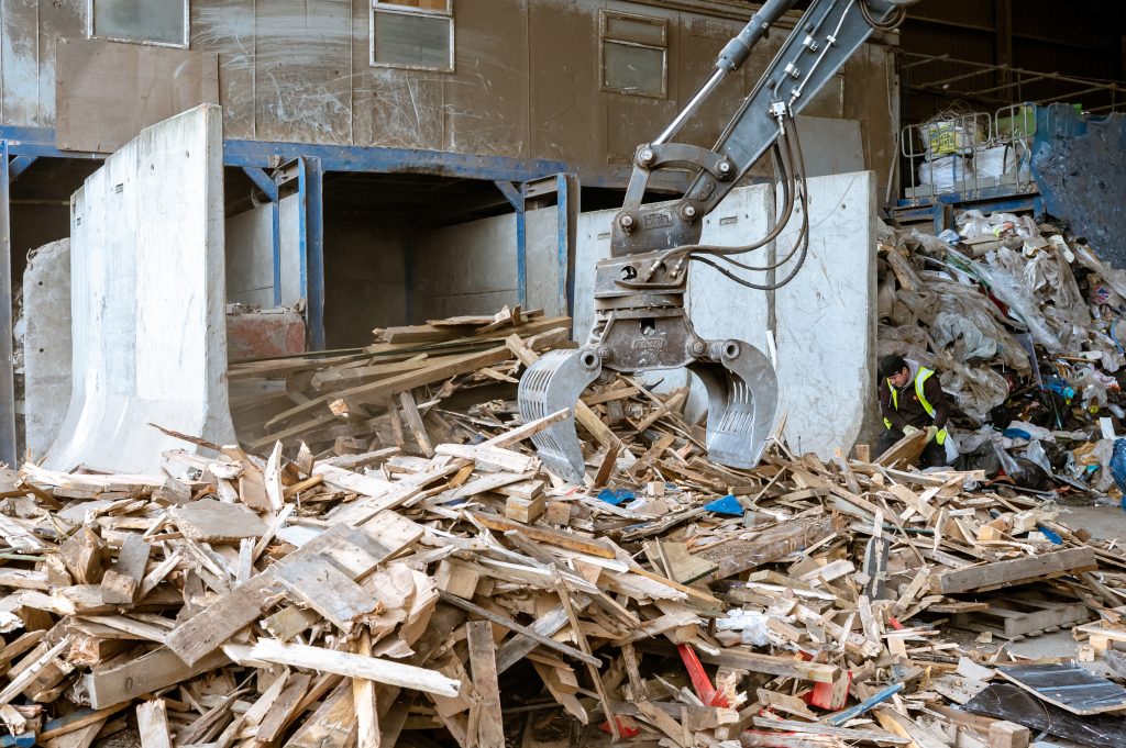 A mechanical claw lifts and moves a pile of wooden debris and waste inside a recycling or waste management facility. Piles of scrap wood and other materials are visible, with a worker in a safety vest nearby.