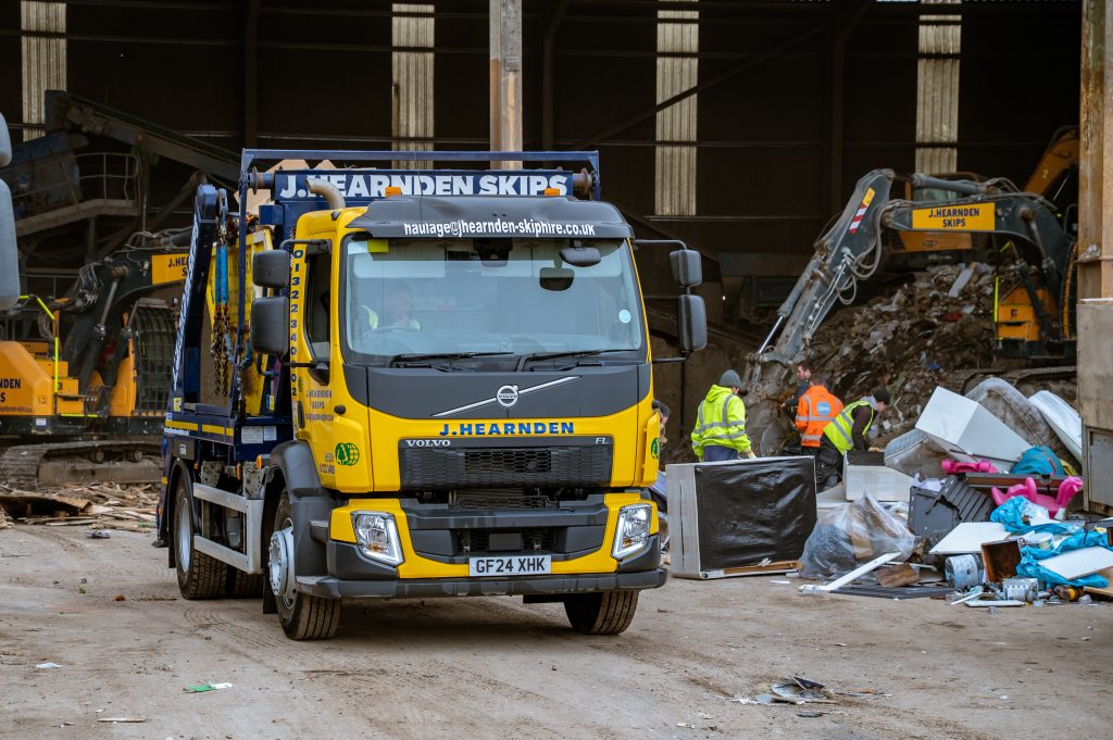 A yellow J. Hearnden Skips lorry is parked inside a large warehouse, surrounded by debris and construction equipment. Two workers in high-visibility clothing are seen sorting through waste in the background.