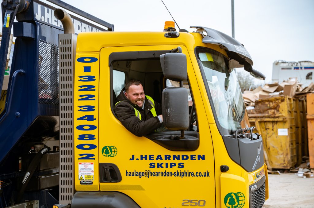 A smiling man wearing a high-visibility jacket sits in the driver’s seat of a yellow J. Hearnden Skips lorry, surrounded by industrial skips and piles of materials in a work yard.