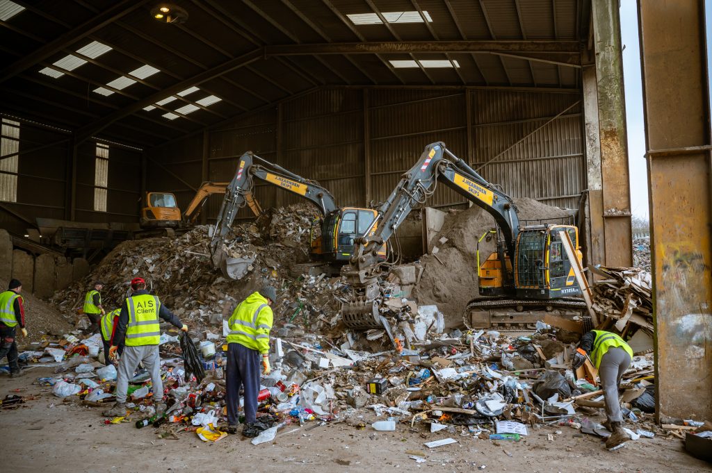 Workers in high-visibility vests sort through piles of waste inside a large industrial building, while excavators move and manage the rubbish in the background.