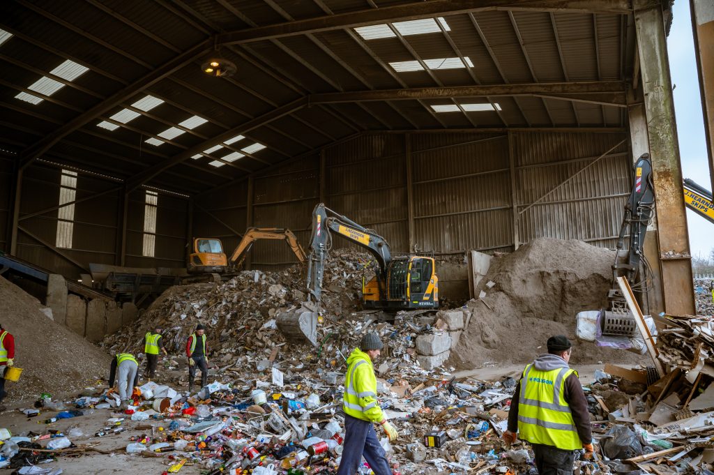 Workers in high-visibility vests sort through large piles of waste and recyclables inside a warehouse, whilst excavators move materials in the background.