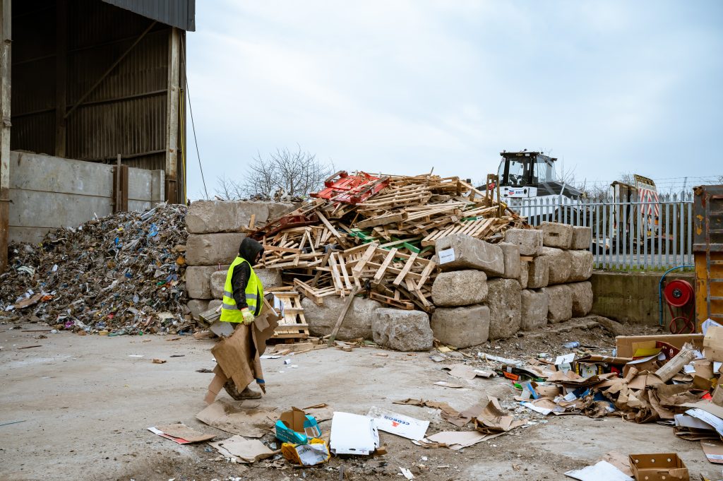 A person in a yellow safety jacket walks near piles of wooden pallets, bales of compressed material, and assorted debris at a recycling or waste management facility. A tractor is visible in the background.