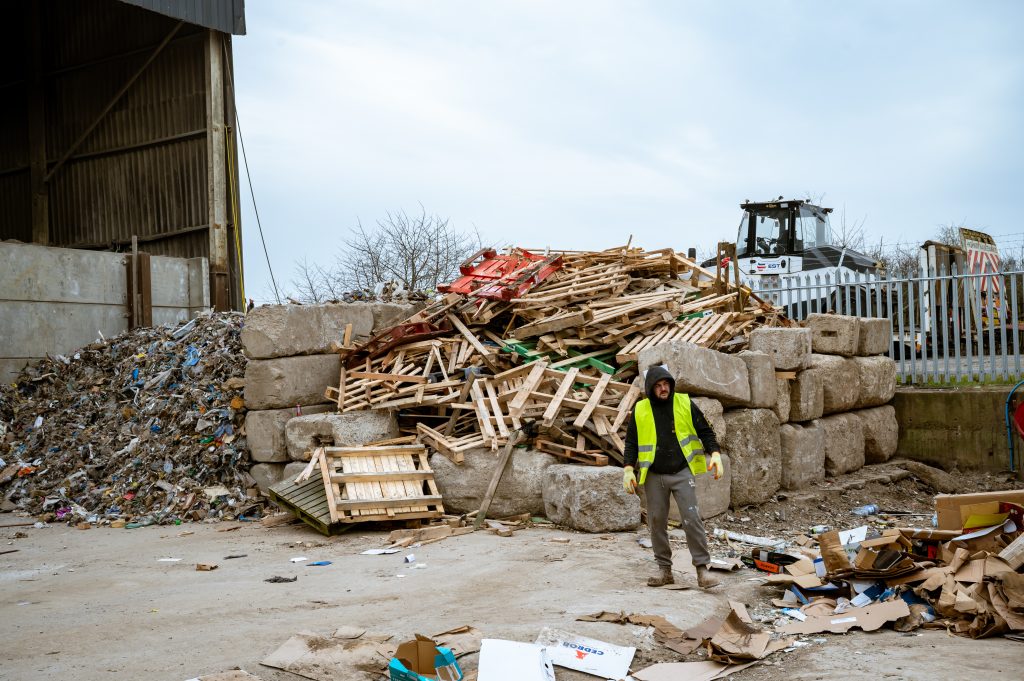 A worker in a yellow safety vest stands near a large pile of wooden pallets and debris at an industrial waste site, with a tractor and metal fence in the background.