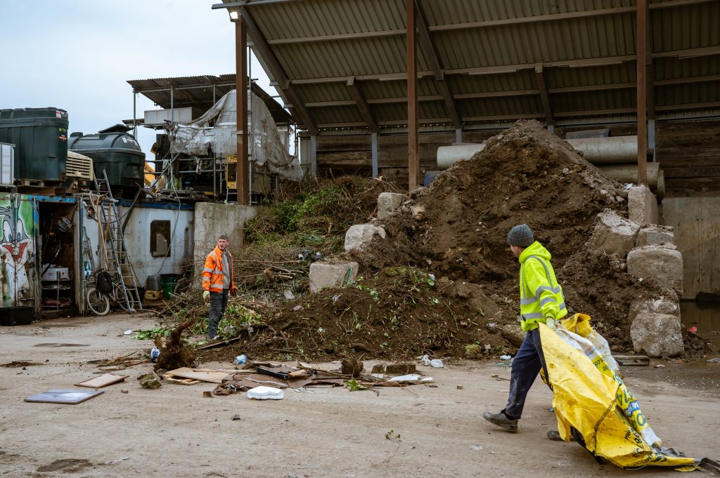 Two workers in high-visibility jackets move debris and a large yellow bag at an outdoor construction or waste site, with piles of earth, scattered rubbish, and industrial equipment in the background.