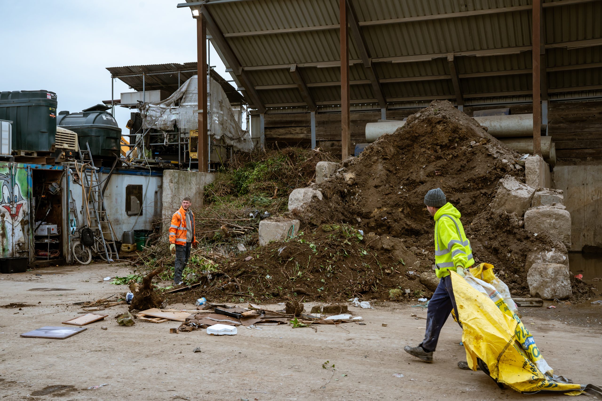 Two workers in high-visibility jackets move debris and a large yellow bag at an outdoor construction or waste site, with piles of earth, scattered rubbish, and industrial equipment in the background.