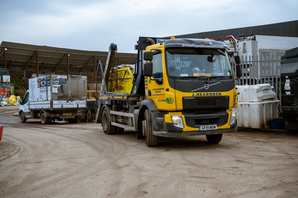 A yellow J. Hearnden Volvo skip lorry is parked at a construction or industrial yard, with a skip loaded on the back. Other vehicles and equipment are visible in the background, along with a large shed.