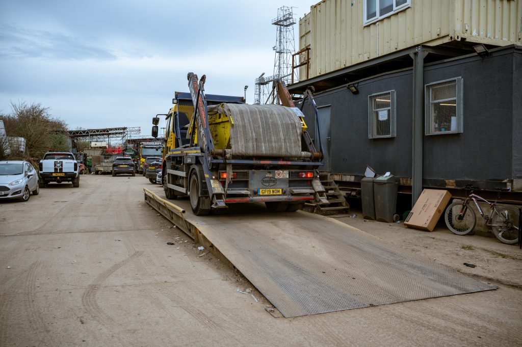 A lorry carrying a large metal drum is parked on a metal ramp beside a black building in an industrial yard, with other vehicles and construction materials visible in the background.