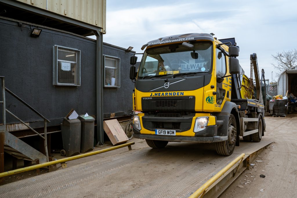 A yellow Volvo skip lorry with "J. Hearnden" branding is parked on a weighbridge beside black portable buildings at an industrial site; bins and rubbish are visible nearby.