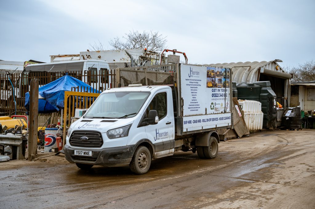 A white flatbed lorry with a company sign on the side is parked in a muddy industrial yard, surrounded by various equipment, metal fencing, and temporary structures under an overcast sky.