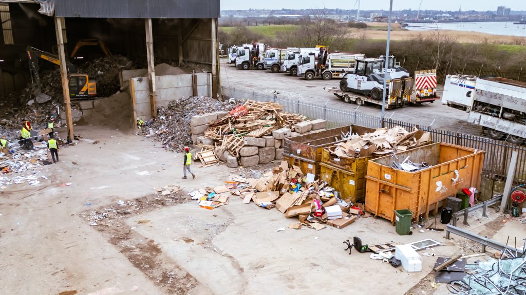 A recycling centre with workers sorting waste, large piles of debris, containers filled with wood and scrap, and several lorries parked near an open building. The area overlooks a body of water and distant buildings.