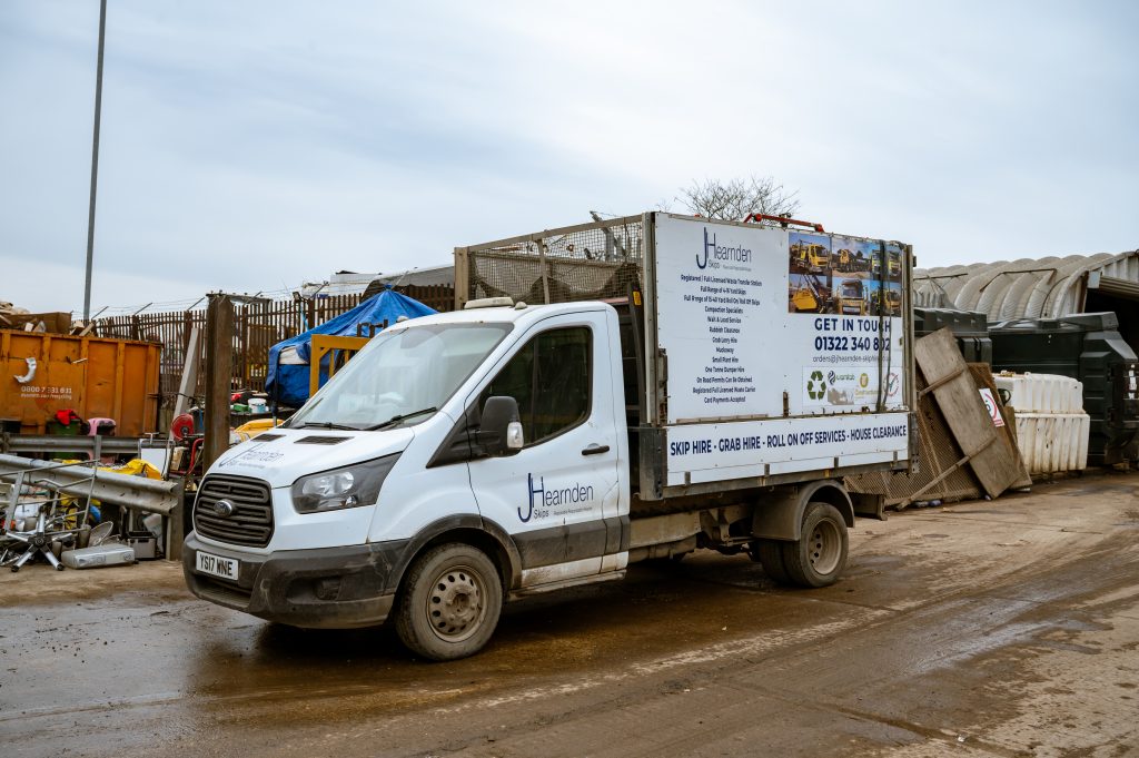 A white Hernden Waste van is parked in a muddy industrial yard, surrounded by skips, bins, and building materials. The van displays contact information and a list of waste removal services on its side panel.