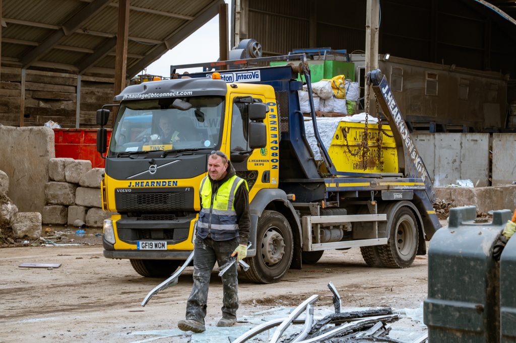 A worker in a high-visibility vest stands in front of a yellow and blue Hernden lorry loaded with scrap materials at an industrial site with metal debris on the ground and large bales in the background.