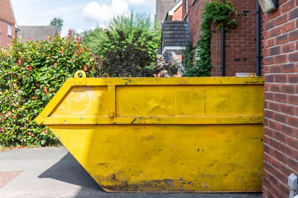 A large yellow metal skip sits on a driveway beside a red brick house, with green bushes and other houses in the background on a sunny day.