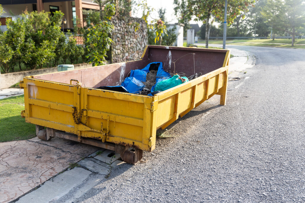 A large yellow skip filled with bags of rubbish and debris sits on the edge of a residential street near a pavement and houses, with greenery and sunlight in the background.