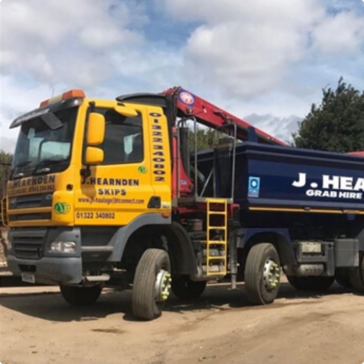 A yellow and blue J. Hearnden Grab Hire lorry is parked on a dirt road under a cloudy sky, with contact information displayed on the cab and skip hire equipment visible.