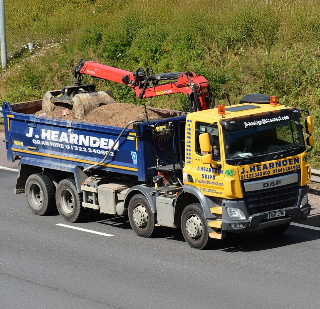 A blue and yellow J. Hearnden tipper lorry with a grabber arm is driving on a road, carrying large rocks and soil in its open cargo bed. The lorry has company details on the side and is on a clear, sunny day.