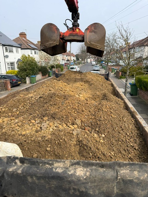 A mechanical digger with a red scoop is unloading a large pile of soil and rocks onto a lorry bed parked on a residential street lined with houses and cars.