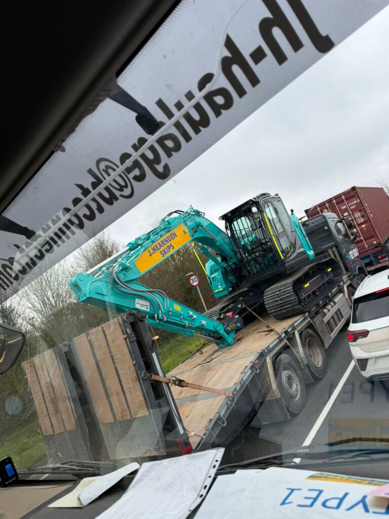 A teal and yellow excavator labelled "JARCOHENSE SITES" is being transported on a flatbed lorry in heavy traffic, viewed from inside another vehicle. Other cars and shipping containers are visible on the road.